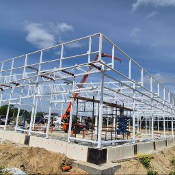 LEKSTAL KONSTRUKCJE - Biała konstrukcja stalowa hali w trakcie budowy, widoczny żuraw i rusztowania, błękitne niebo z chmurami w tle. Fundamenty betonowe z izolacją.