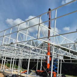 LEKSTAL KONSTRUKCJE - Konstrukcja stalowa hali w trakcie montażu, widoczny dźwig i pracownicy. Białe profile na tle błękitnego nieba z chmurami. Częściowo zabezpieczona folią.