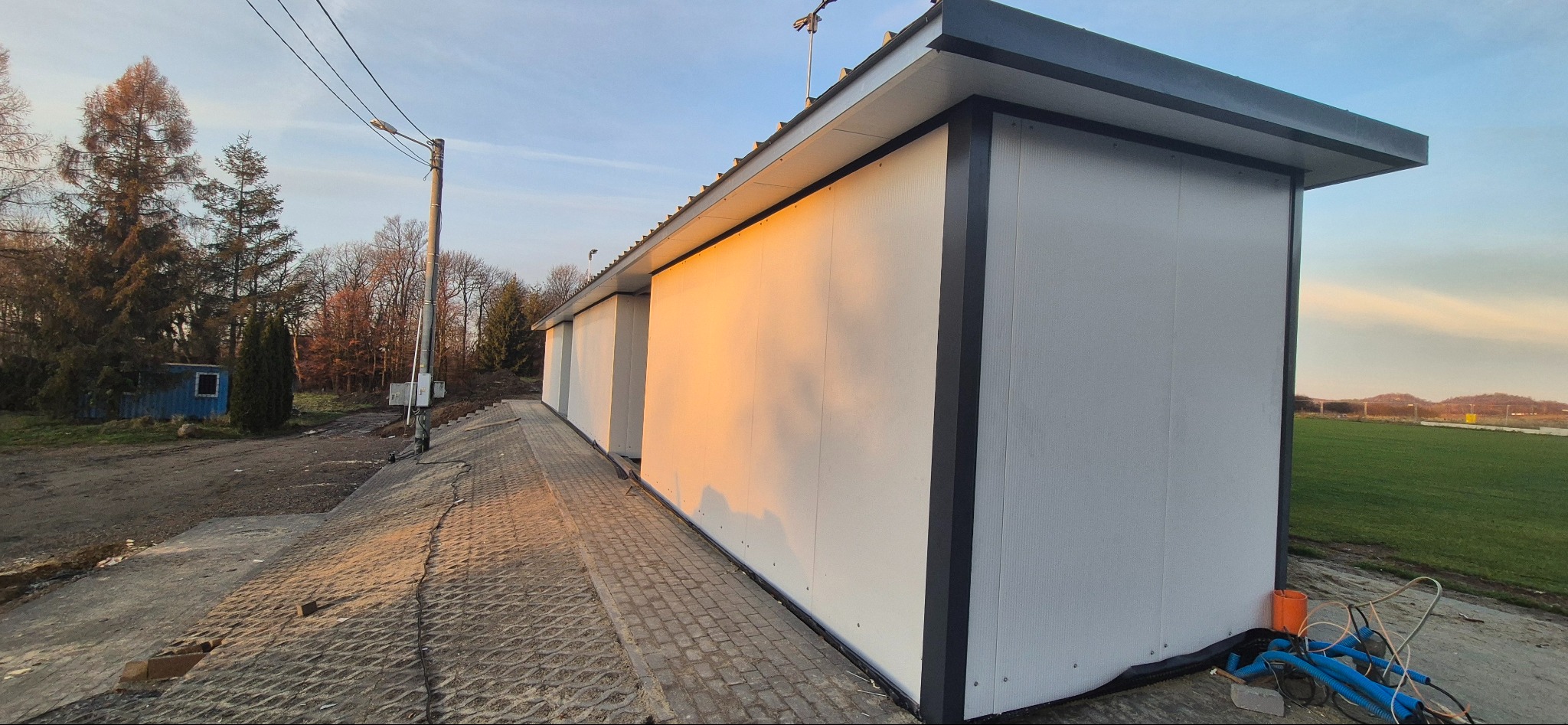 Hala z płyt warstwowych w promieniach zachodzącego słońca, widok z boku, na pierwszym planie chodnik z kostki brukowej, w tle drzewa i łąka.