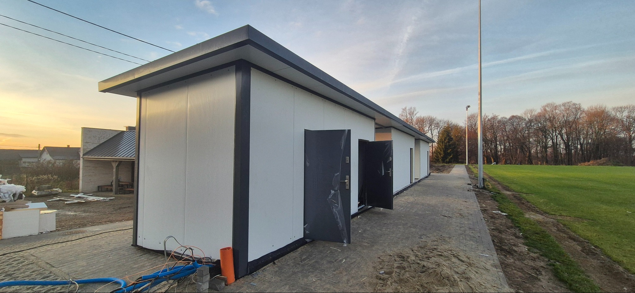 Nowoczesny pawilon z płyt warstwowych z otwartymi drzwiami, widok z zewnątrz, oświetlony zachodzącym słońcem. Widoczne kable i otoczenie budowy.