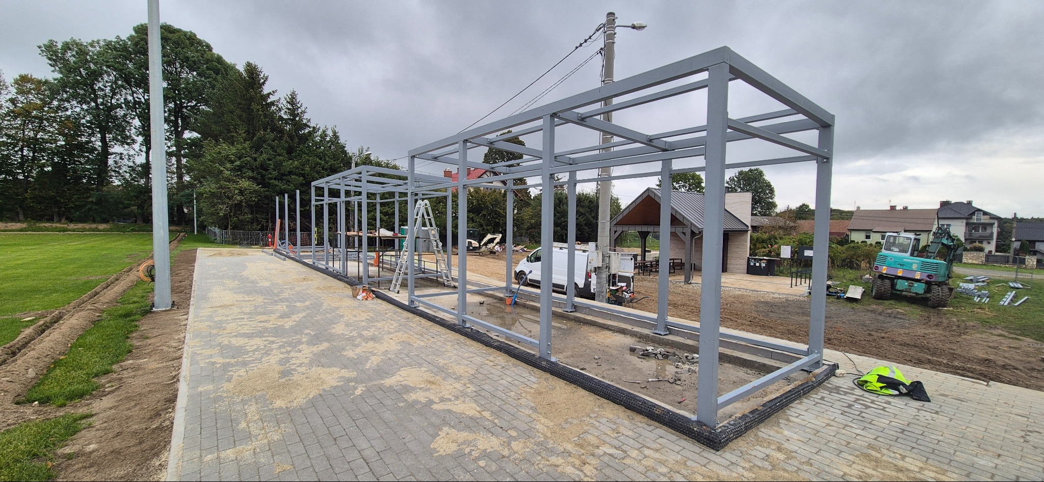 Szara, metalowa konstrukcja z drabinką w tle, prawdopodobnie zadaszenie przystanku autobusowego, w trakcie budowy na tle zieleni i zabudowań.