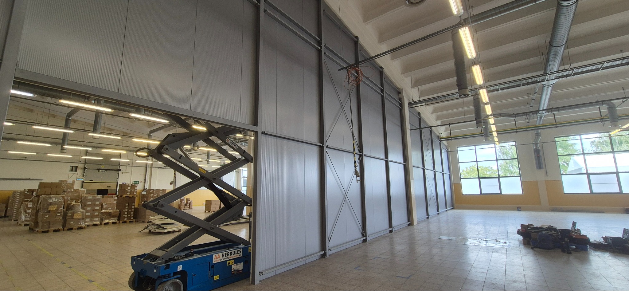 Wnętrze hali w trakcie montażu płyt warstwowych, widoczny podnośnik nożycowy i częściowo zamontowana ściana z płyt. Oświetlenie przemysłowe, posadzka z płytek.