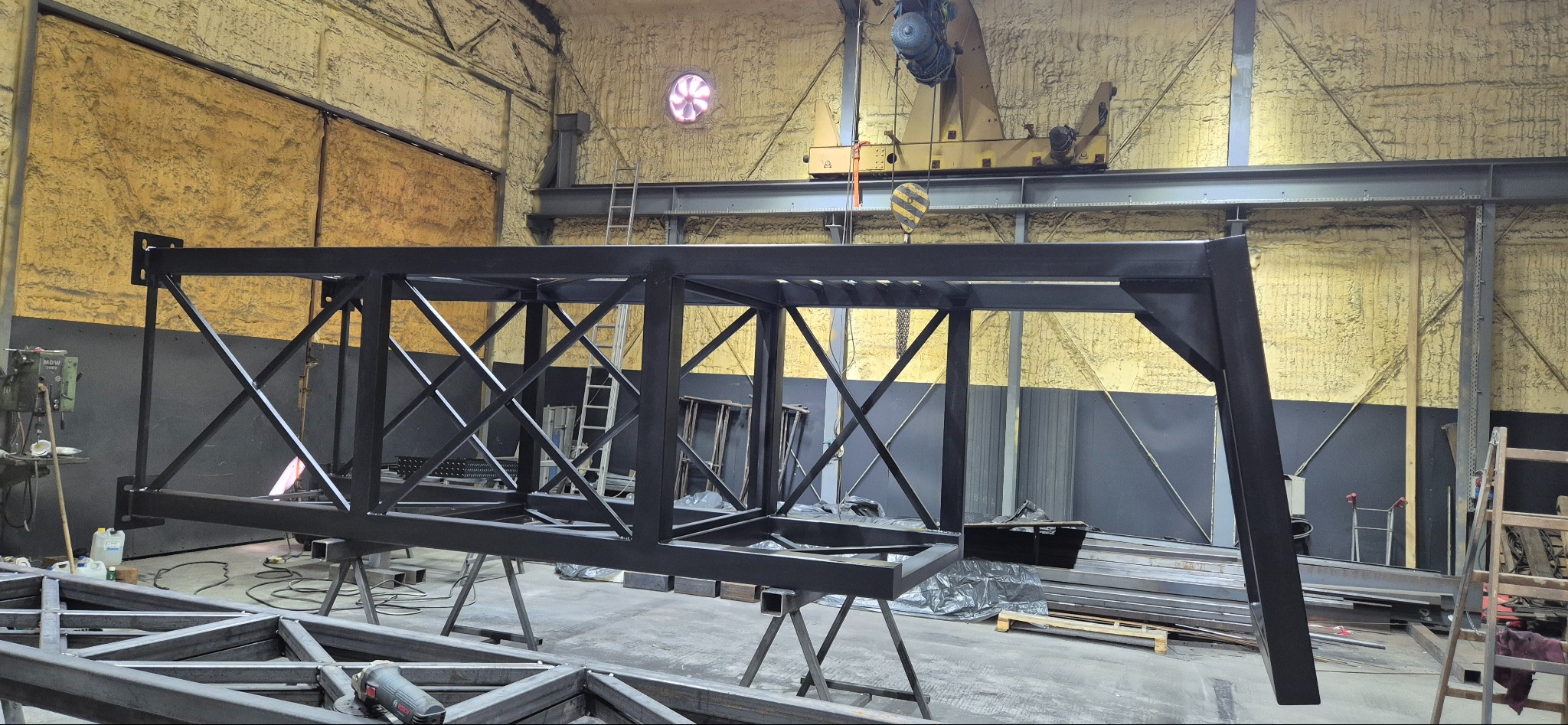 Czarna, stalowa konstrukcja kratowa w hali produkcyjnej z żółtymi ścianami i suwnicą. Element hali lub mostu w trakcie budowy.