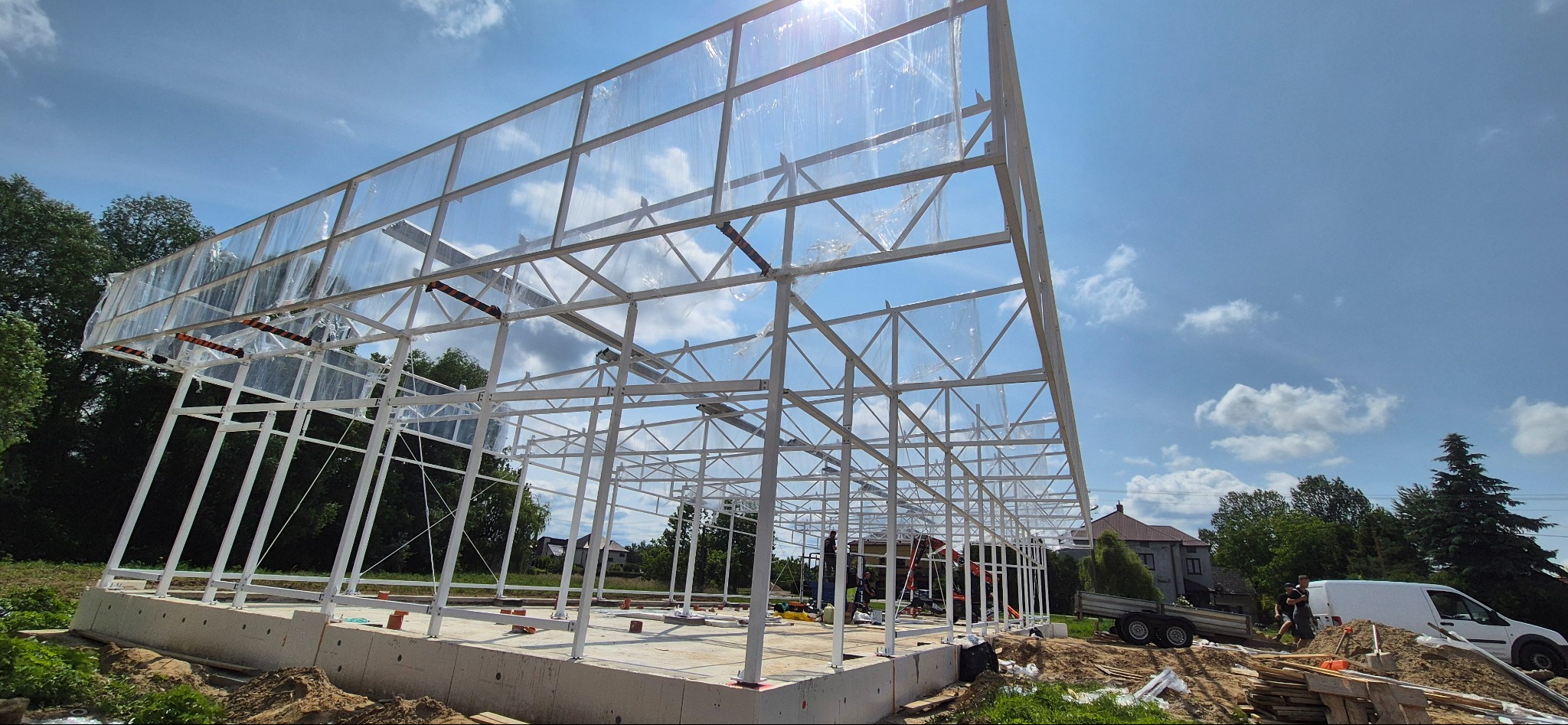 Konstrukcja stalowa hali w trakcie budowy, widok z zewnątrz, białe profile, częściowo pokryte folią, fundament betonowy, błękitne niebo w tle.
