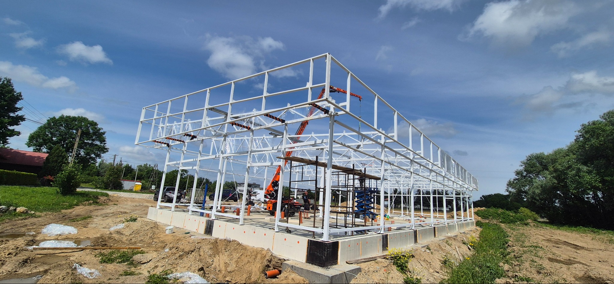 Biała konstrukcja stalowa hali w trakcie budowy, widoczny żuraw i rusztowania, błękitne niebo z chmurami w tle. Fundamenty betonowe z izolacją.