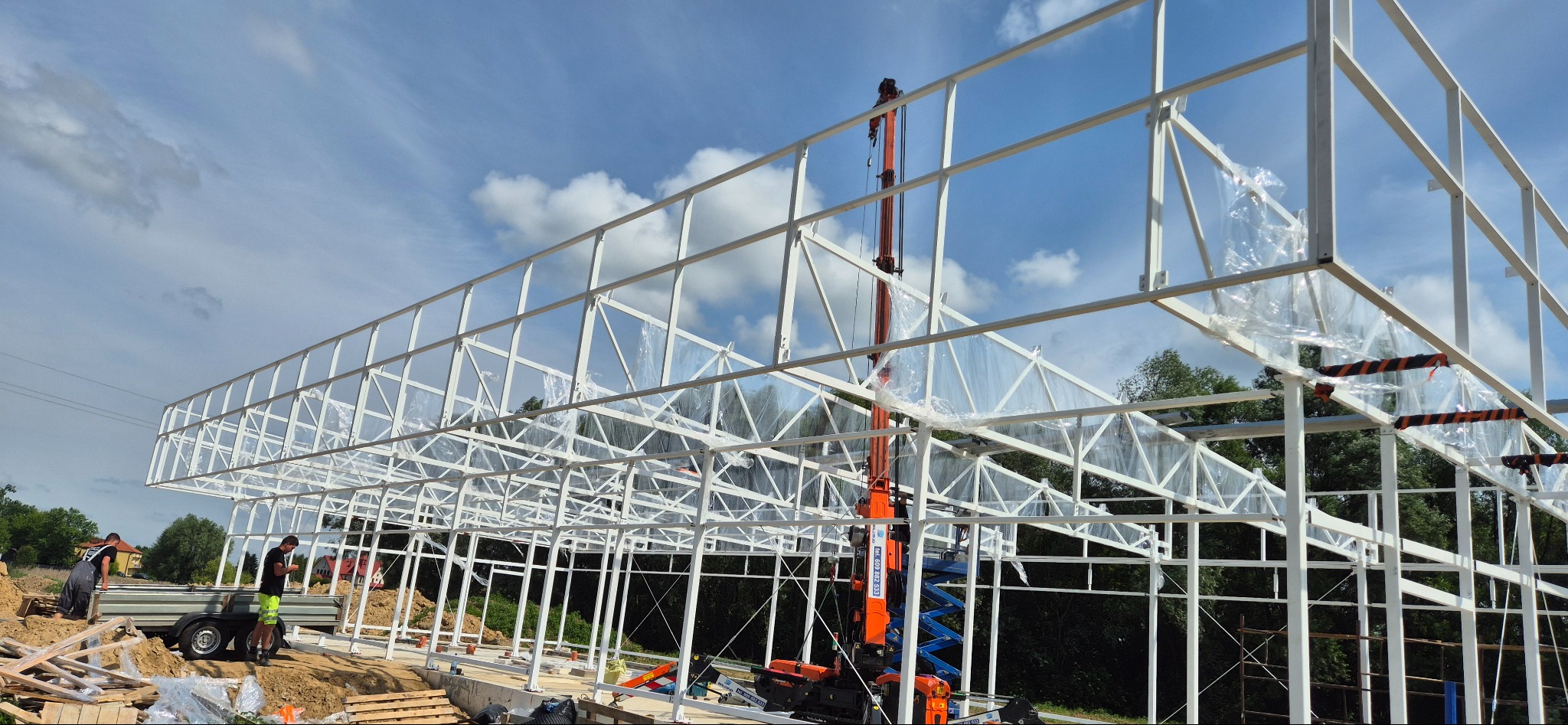 Konstrukcja stalowa hali w trakcie montażu, widoczny dźwig i pracownicy. Białe profile na tle błękitnego nieba z chmurami. Częściowo zabezpieczona folią.