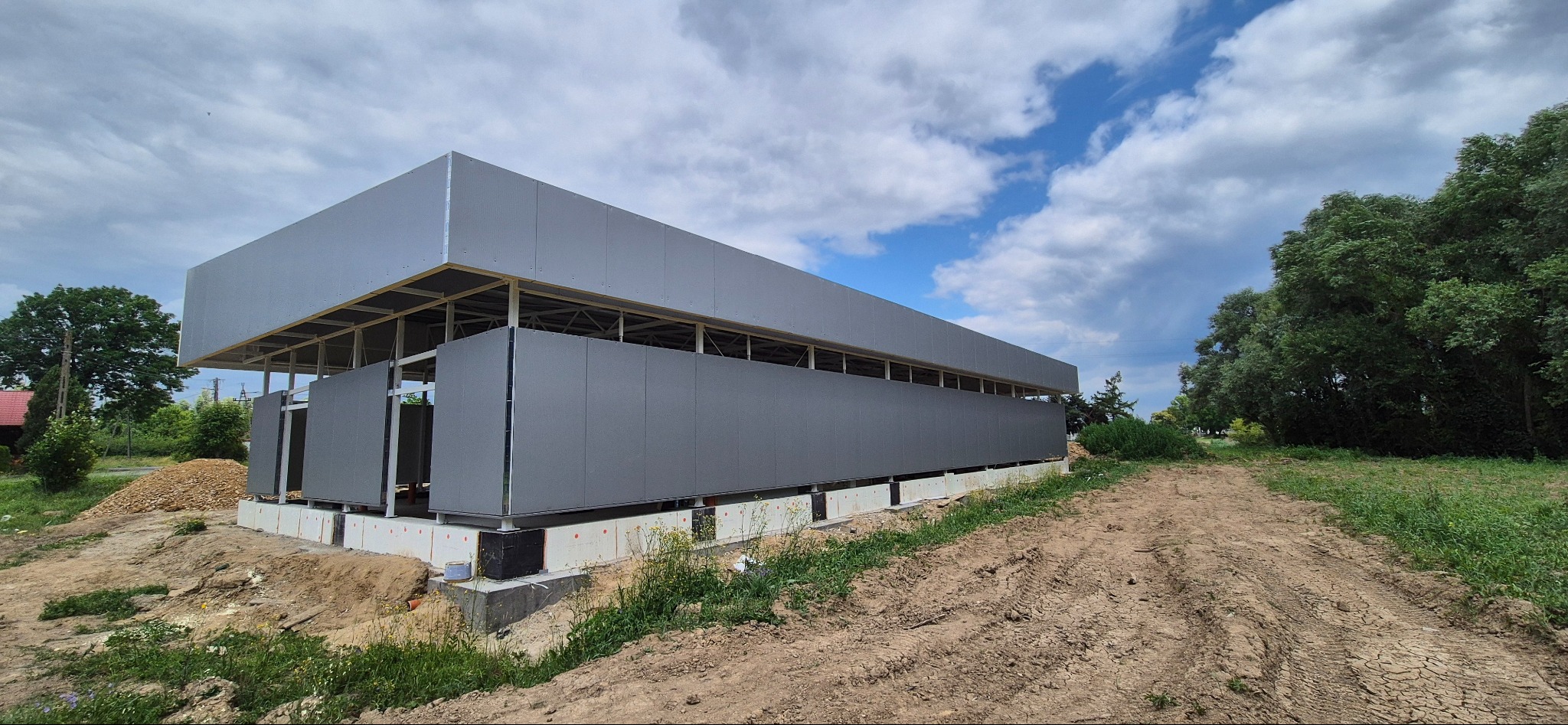 Hala w trakcie budowy, widoczne szare płyty warstwowe i stalowa konstrukcja. W tle błękitne niebo z chmurami i zieleń drzew. Ujęcie z poziomu gruntu.