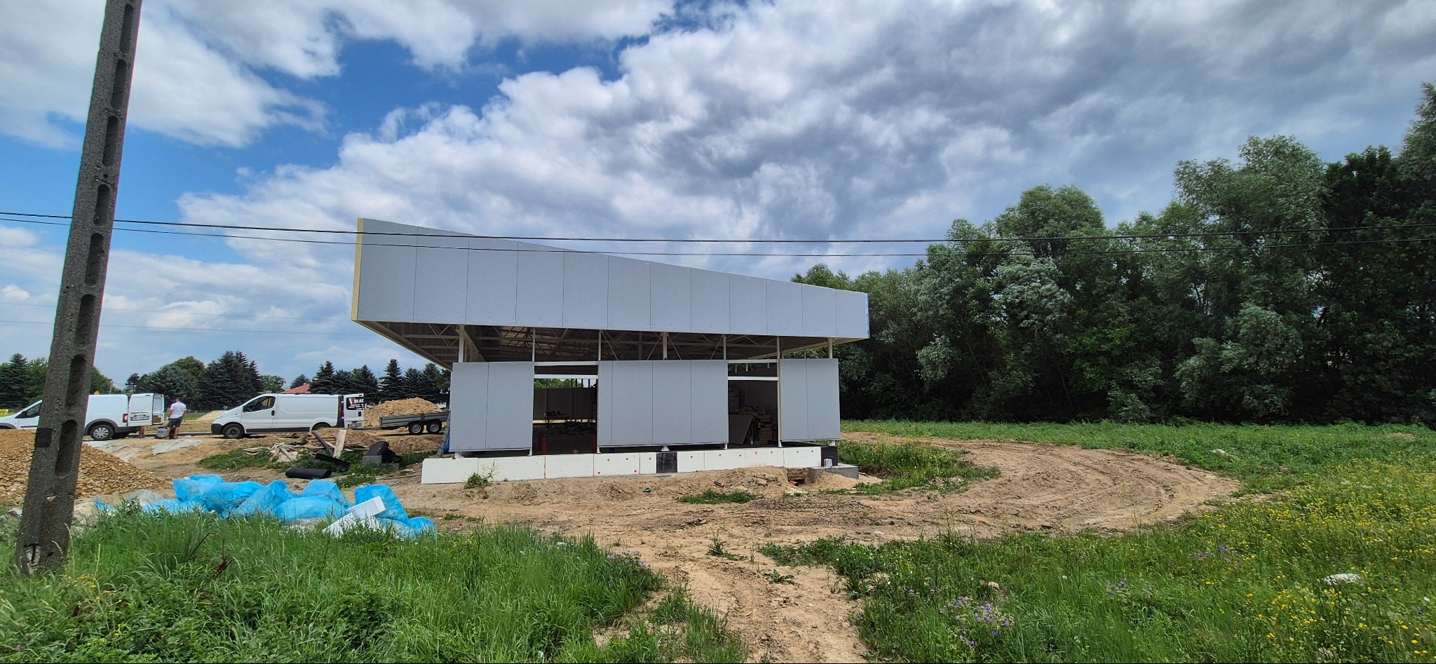 Budowa hali z płyt warstwowych w trakcie realizacji. Widoczne elementy konstrukcyjne, teren budowy i pojazdy. Nietypowy kształt dachu.