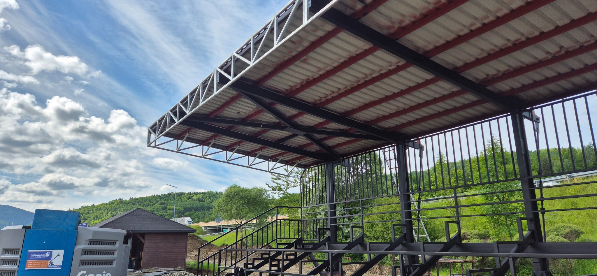 Zadaszona konstrukcja stalowa z metalowymi schodami na tle zieleni i błękitnego nieba. Widoczna blacha trapezowa i elementy konstrukcyjne.