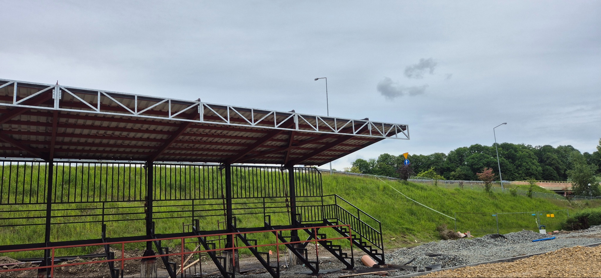 Zadaszona konstrukcja stalowa z metalowymi schodami na tle zielonego wzgórza i pochmurnego nieba. Widoczne elementy konstrukcyjne i otoczenie placu budowy.