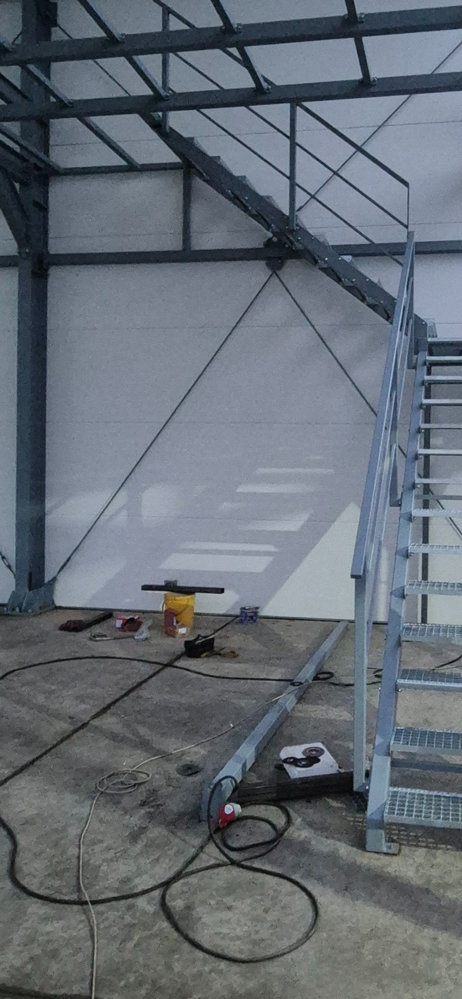 Wnętrze hali w trakcie budowy: widoczne metalowe schody, konstrukcja dachu, ściany z płyt warstwowych i narzędzia na podłodze. Ujęcie z dołu, oświetlenie robocze.