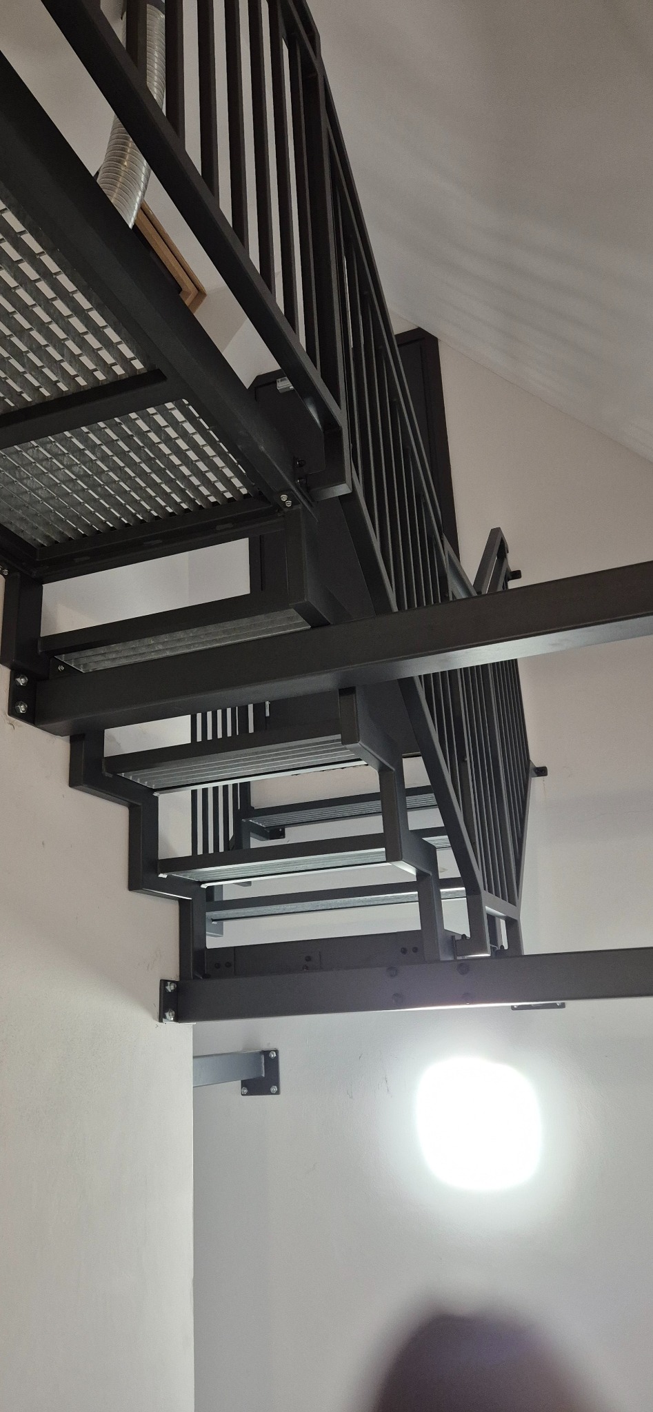 Nowoczesne, czarne schody metalowe z ażurowymi stopniami i minimalistycznymi barierkami, zamontowane w jasnym wnętrzu. Widok z dołu, podkreślający konstrukcję.