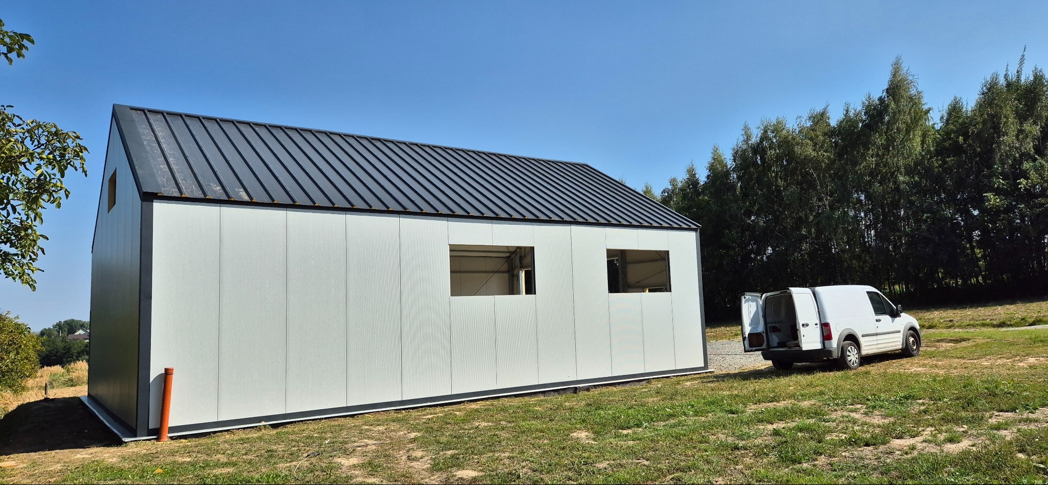 Gotowa hala z płyt warstwowych z otworami okiennymi, na tle zieleni i błękitnego nieba. Biały bus z otwartymi drzwiami stoi obok.