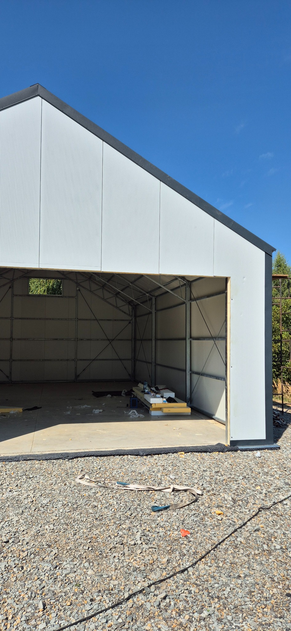 Wnętrze hali w trakcie budowy, widoczne płyty warstwowe i stalowa konstrukcja. Na zewnątrz gruz i błękitne niebo. Hala z widocznym oknem.