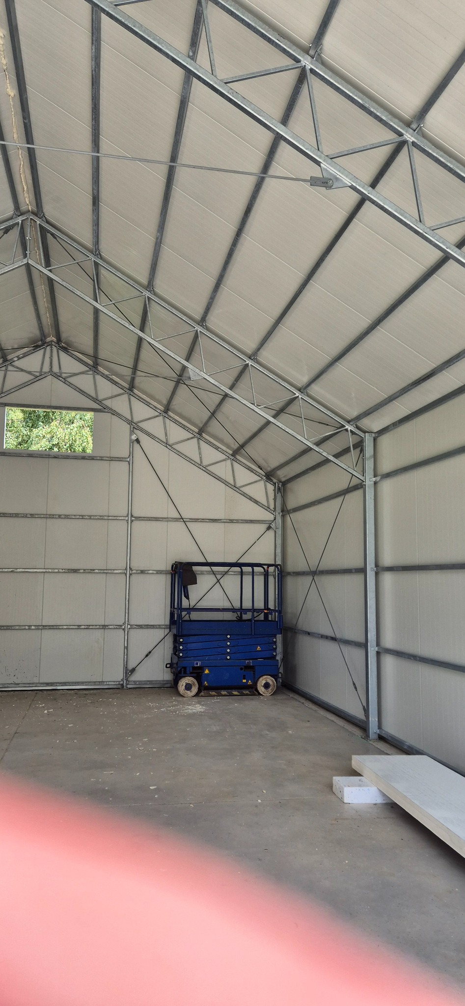 Wnętrze hali z metalową konstrukcją i płytami warstwowymi. Widoczny podnośnik koszowy i okno z zielenią w tle. Hala w trakcie budowy, surowy beton na podłodze.