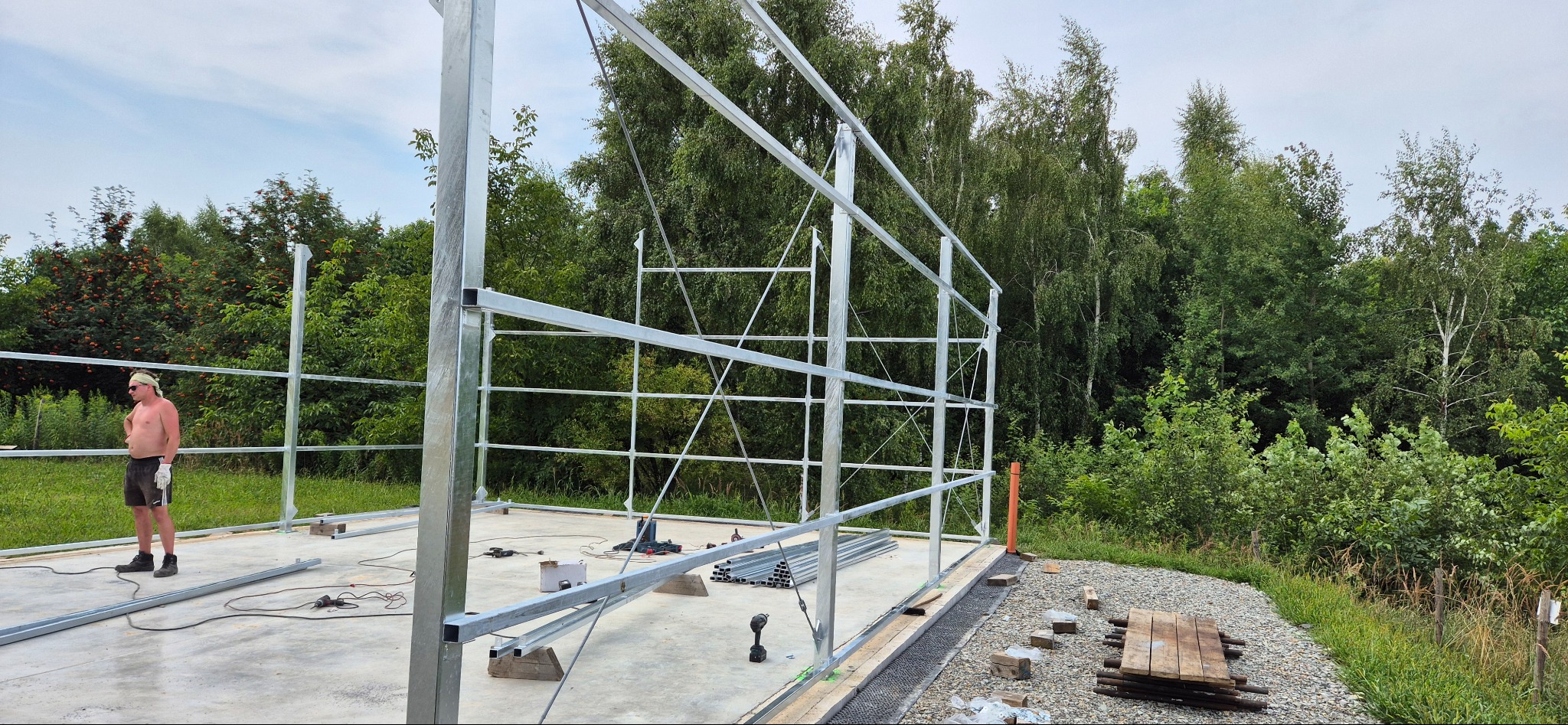 Wznoszenie konstrukcji stalowej hali na betonowej płycie fundamentowej. Widoczny pracownik i narzędzia. Tło stanowią drzewa i krzewy.