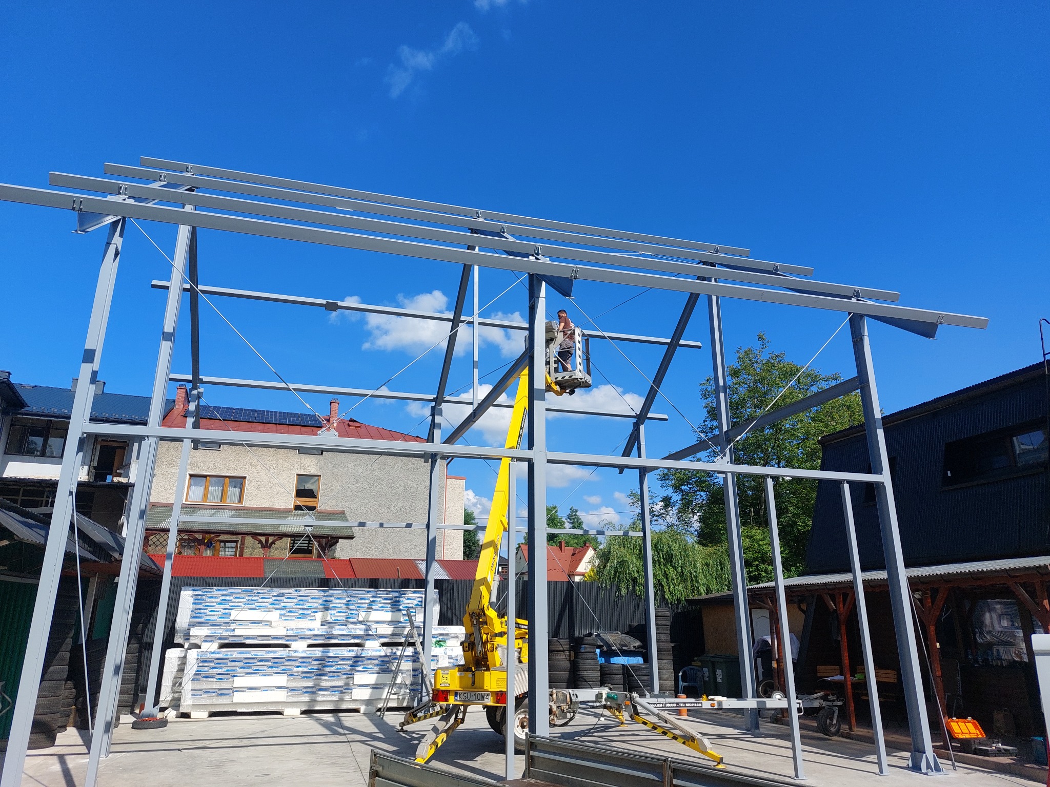 Montaż stalowej konstrukcji hali z użyciem podnośnika koszowego na tle błękitnego nieba i zabudowań. Widoczne płyty warstwowe.