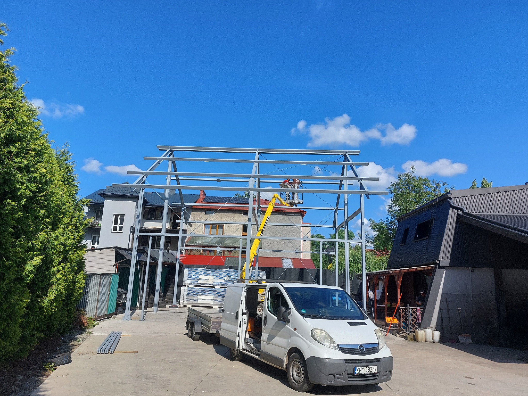 Montaż szkieletu hali stalowej z podnośnika koszowego na tle błękitnego nieba. Na pierwszym planie biały bus z otwartymi drzwiami i przyczepką.