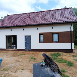 Usługi ciesielsko budowlane - Dom jednorodzinny z nowym, bordowym dachem z dachówki ceramicznej, białą elewacją z brązowymi akcentami, widoczne przygotowania do prac wykończeniowych wokół budynku.