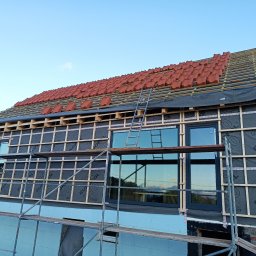 Usługi ciesielsko budowlane - Dom w trakcie budowy dachu z widoczną konstrukcją więźby, membraną dachową, częściowo ułożoną czerwoną dachówką, rusztowaniami i oknami.