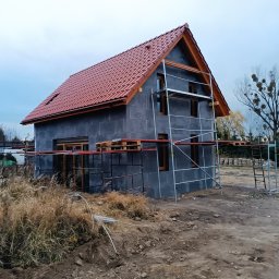 Usługi ciesielsko budowlane - Surowy dom jednorodzinny w trakcie budowy, z czerwoną dachówką, szarymi ścianami i drewnianą stolarką okienną, otoczony rusztowaniami i niezagospodarowanym terenem.