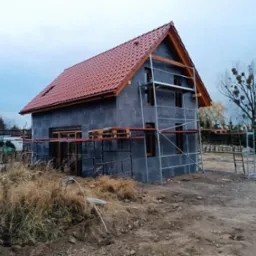 Surowy dom jednorodzinny w trakcie budowy, z czerwoną dachówką, szarymi ścianami i drewnianą stolarką okienną, otoczony rusztowaniami i niezagospodarowanym terenem.