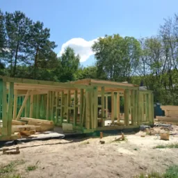 Drewniana konstrukcja szkieletowa domu w trakcie budowy, widoczne pionowe słupki i poziome belki, impregnowane na zielono, na tle drzew i błękitnego nieba z chmurami.