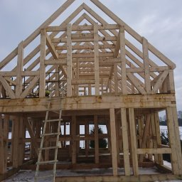 Usługi ciesielsko budowlane - Surowa drewniana konstrukcja domu w trakcie budowy, widoczna więźba dachowa i rusztowanie, zimowy krajobraz w tle.