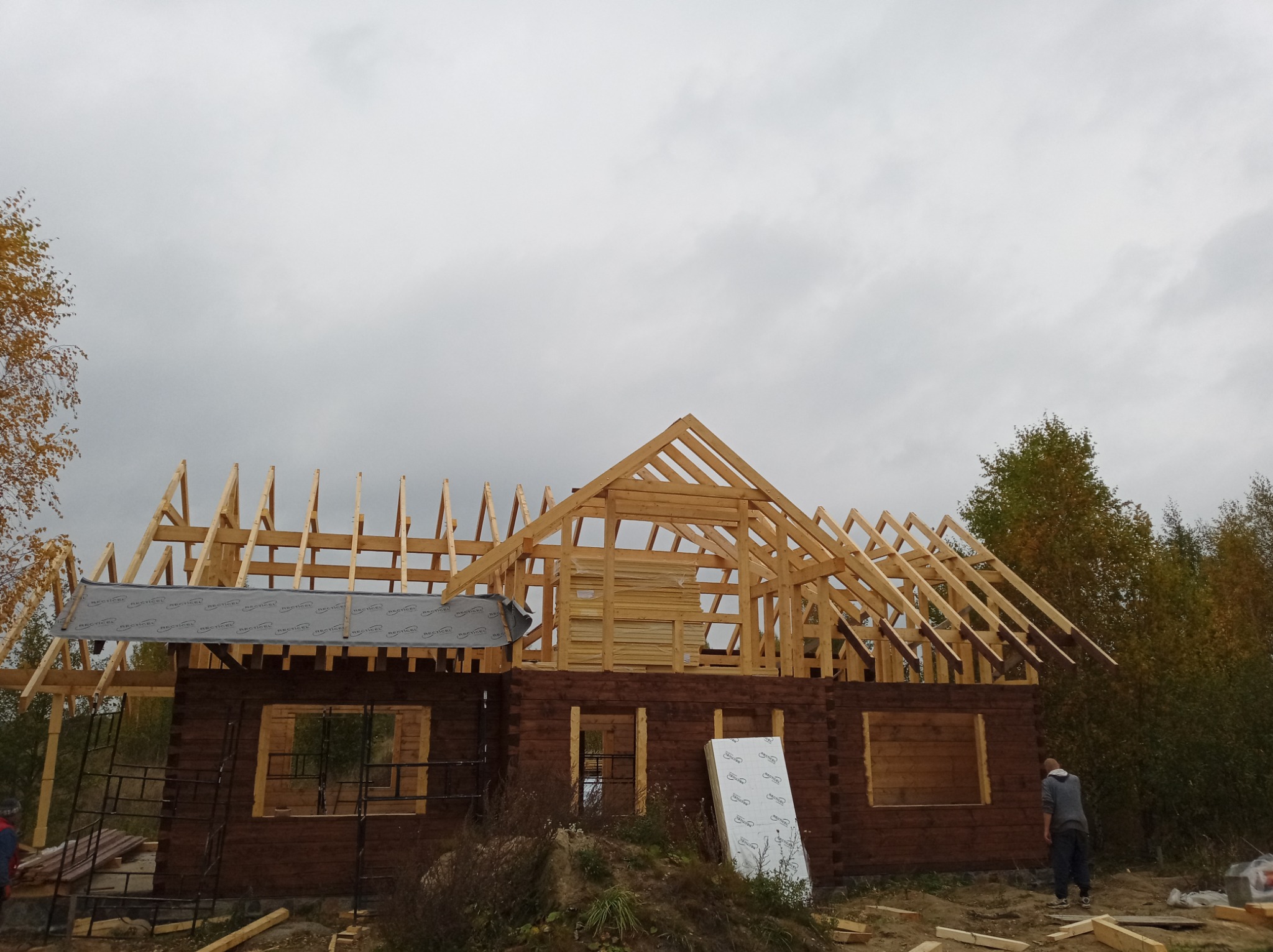 Budowa domu z bali: wznoszenie drewnianej konstrukcji dachu na tle pochmurnego nieba, widoczna osoba przy budynku.