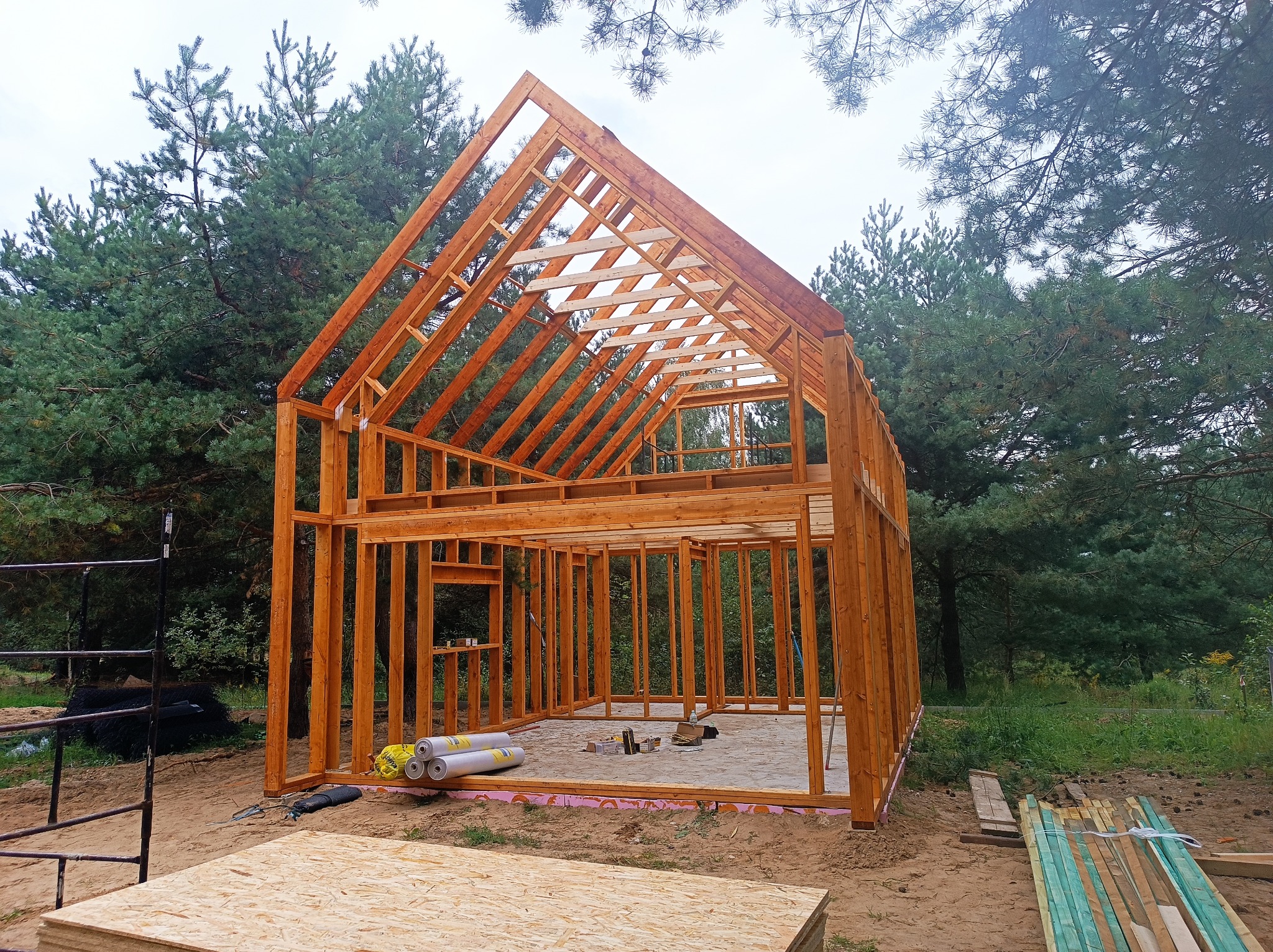 Konstrukcja szkieletowa domu w trakcie budowy, widoczne drewniane belki i słupy, materiały izolacyjne, teren budowy otoczony drzewami.