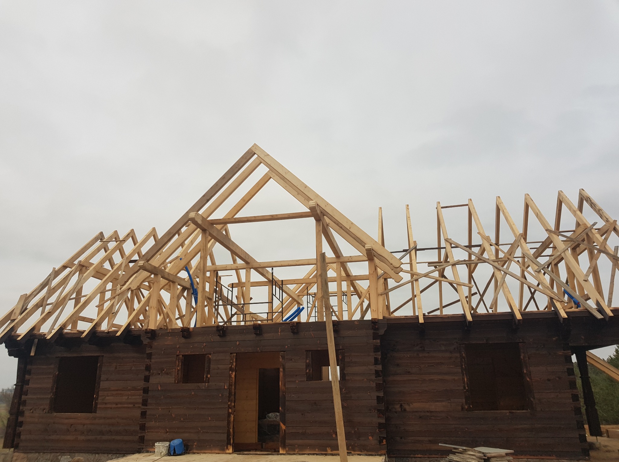 Drewniany dom w trakcie budowy, widoczna konstrukcja ścian z bali i częściowo wykonana drewniana konstrukcja dachu na tle pochmurnego nieba.