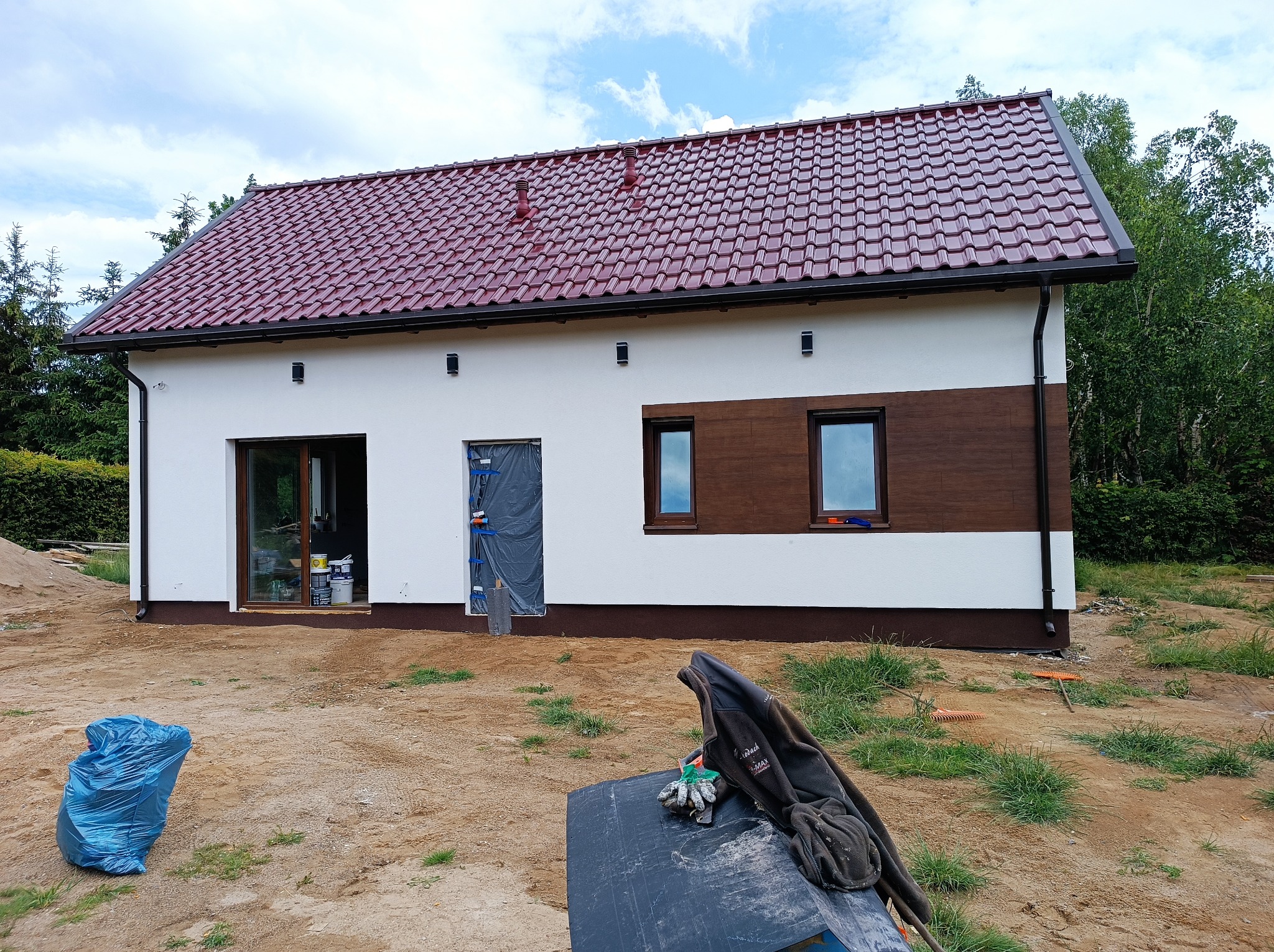 Dom jednorodzinny z nowym, bordowym dachem z dachówki ceramicznej, białą elewacją z brązowymi akcentami, widoczne przygotowania do prac wykończeniowych wokół budynku.