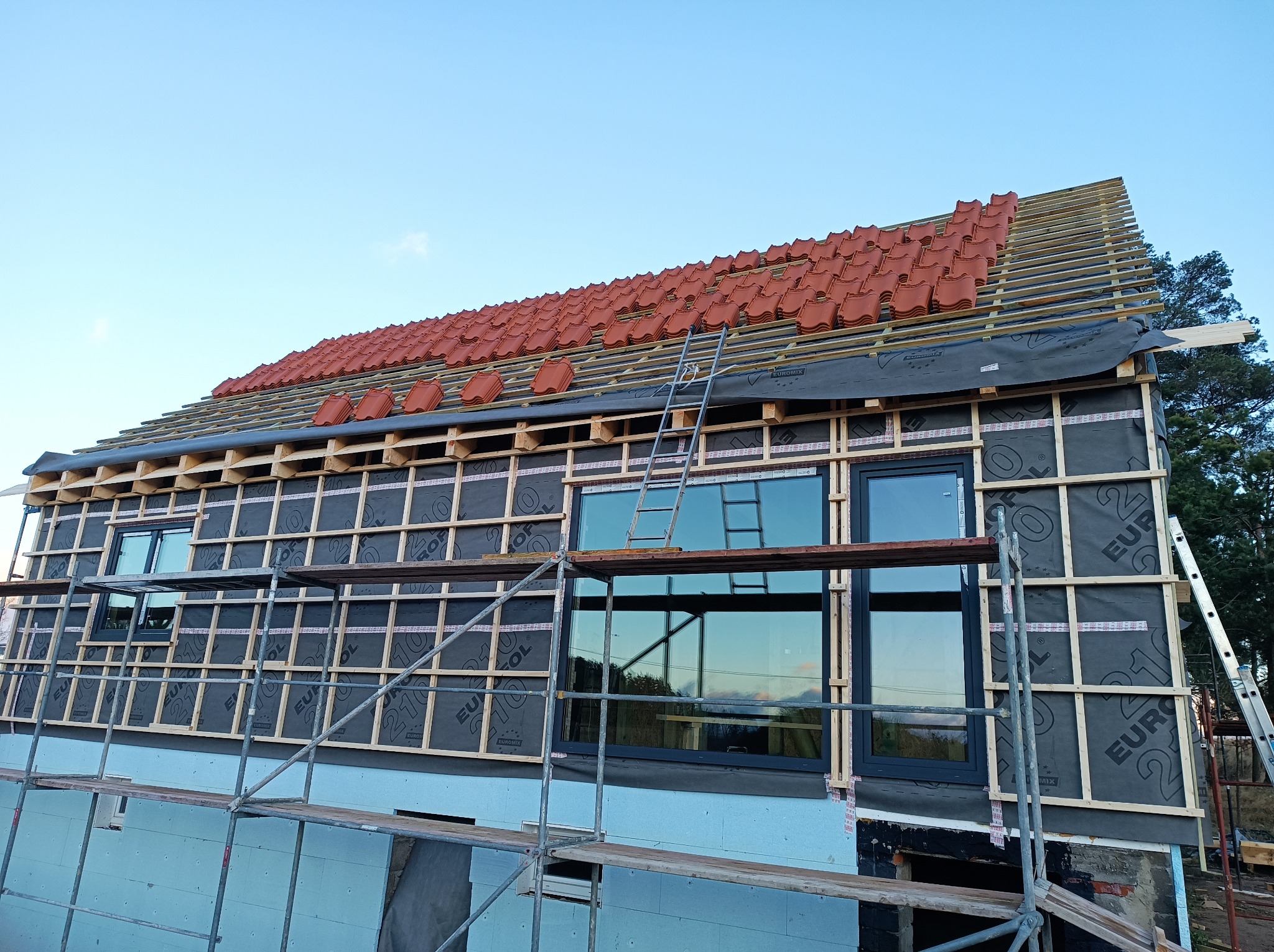 Dom w trakcie budowy dachu z widoczną konstrukcją więźby, membraną dachową, częściowo ułożoną czerwoną dachówką, rusztowaniami i oknami.
