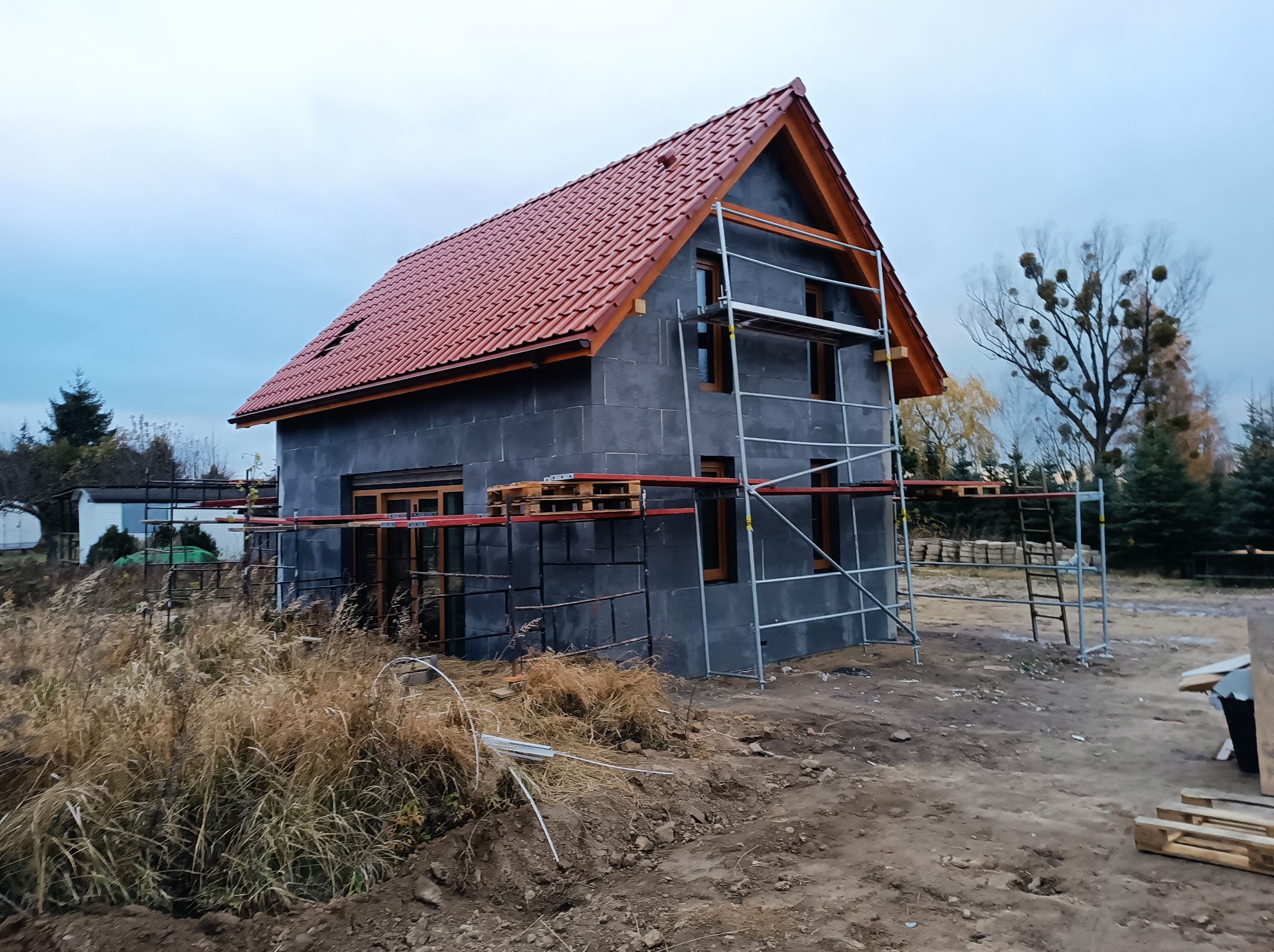 Surowy dom jednorodzinny w trakcie budowy, z czerwoną dachówką, szarymi ścianami i drewnianą stolarką okienną, otoczony rusztowaniami i niezagospodarowanym terenem.