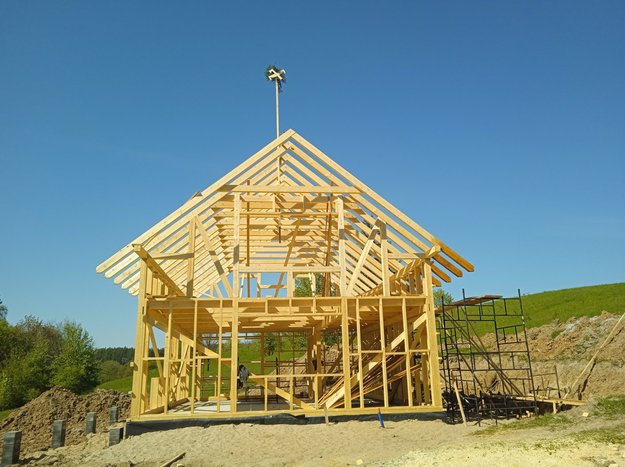 Surowa drewniana konstrukcja domu w trakcie budowy, z wieńcem na szczycie, na tle błękitnego nieba i zieleni.