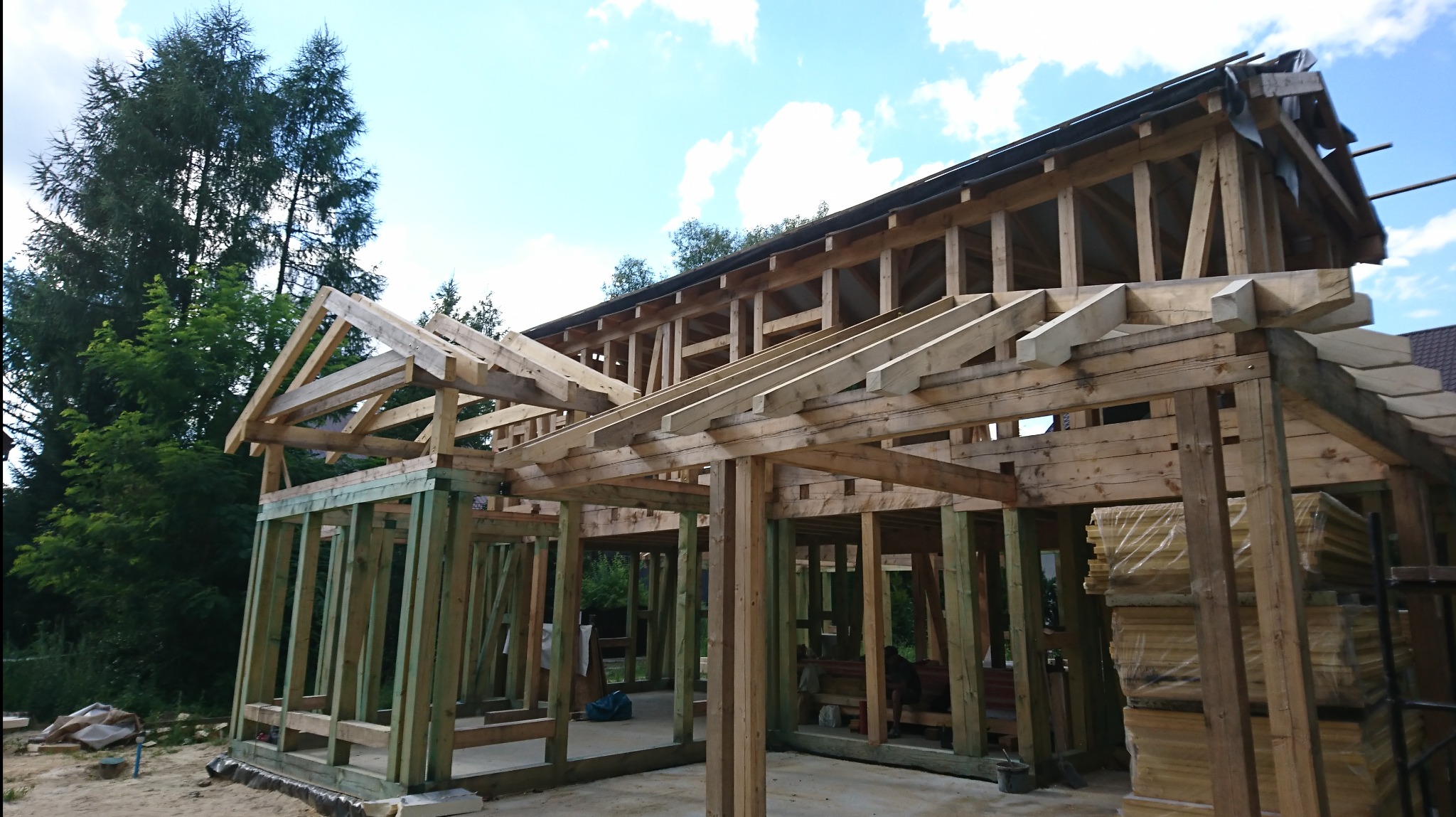 Konstrukcja domu w trakcie budowy z widoczną drewnianą ramą ścian i dachu, częściowo pokrytą czarną folią dachową, na tle zieleni i błękitnego nieba z chmurami.