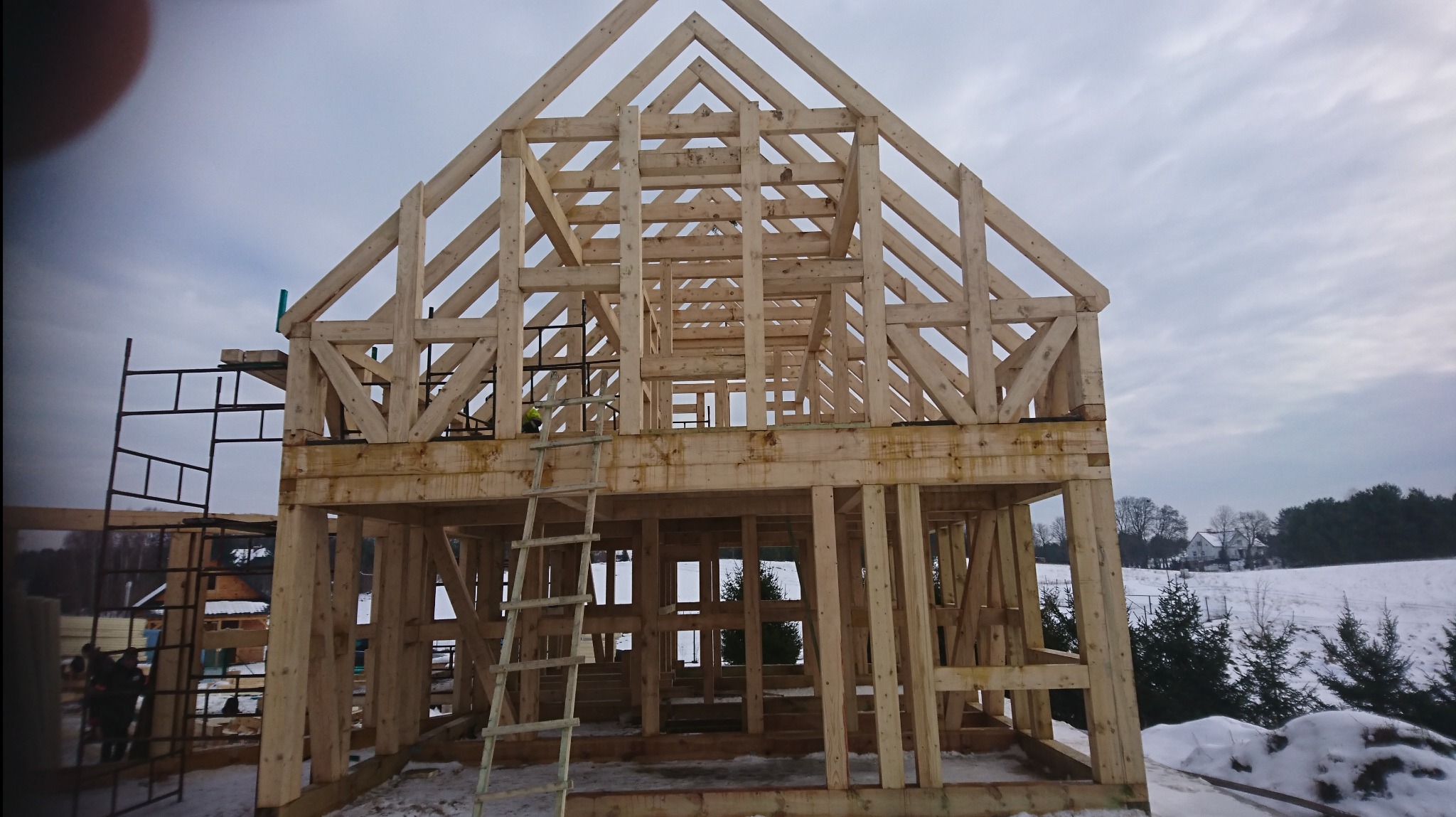 Surowa drewniana konstrukcja domu w trakcie budowy, widoczna więźba dachowa i rusztowanie, zimowy krajobraz w tle.