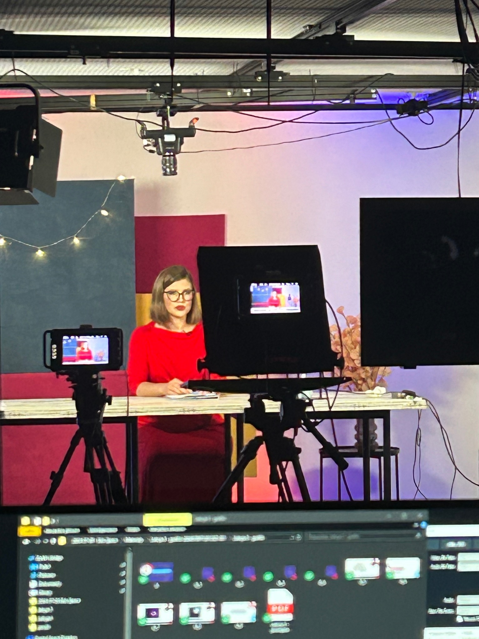 Kobieta w czerwonej sukience i okularach siedzi za stołem w studiu filmowym, otoczona sprzętem do nagrywania, w tym kamerami na statywach i oświetleniem studyjnym.