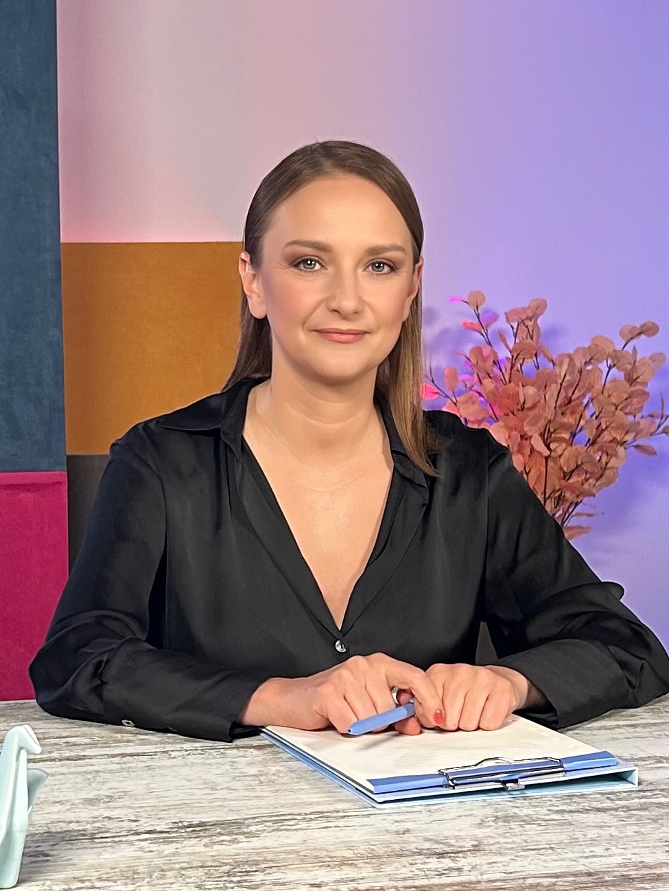 Portret kobiety w czarnej koszuli, siedzącej za biurkiem z niebieskim długopisem i klipsem, z dekoracyjnym tłem w odcieniach różu, fioletu, brązu i zieleni.