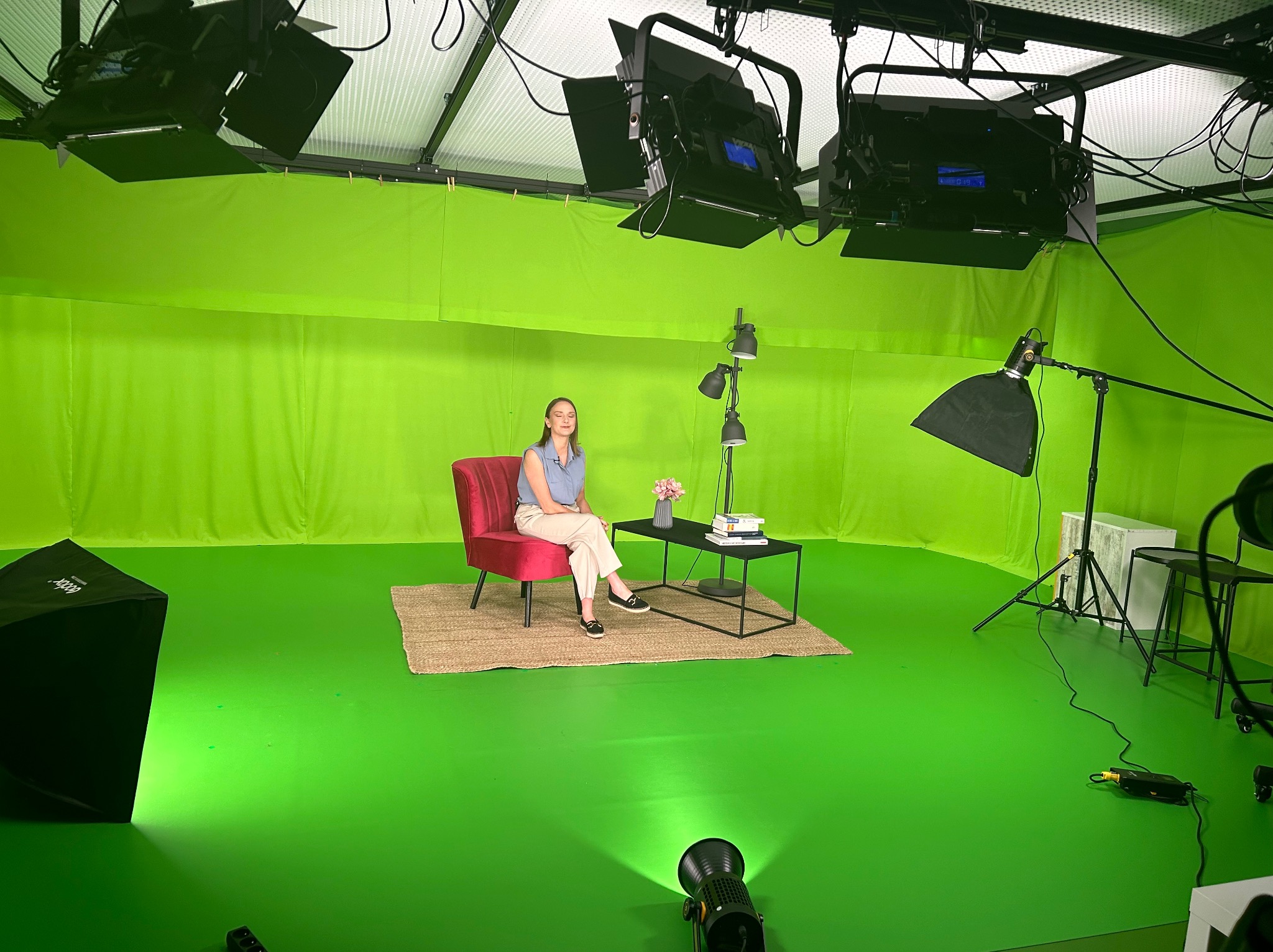 Kobieta siedząca na czerwonym krześle w studio z zielonym tłem, otoczona profesjonalnym sprzętem oświetleniowym i stolikiem kawowym z książkami i kwiatami.