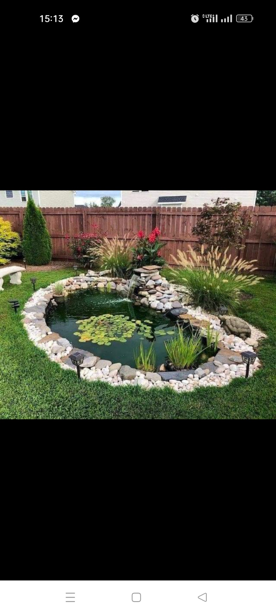 Oczko wodne w ogrodzie, otoczone białymi kamieniami i roślinnością, mały wodospad z kamieni, w tle drewniany płot i trawnik.