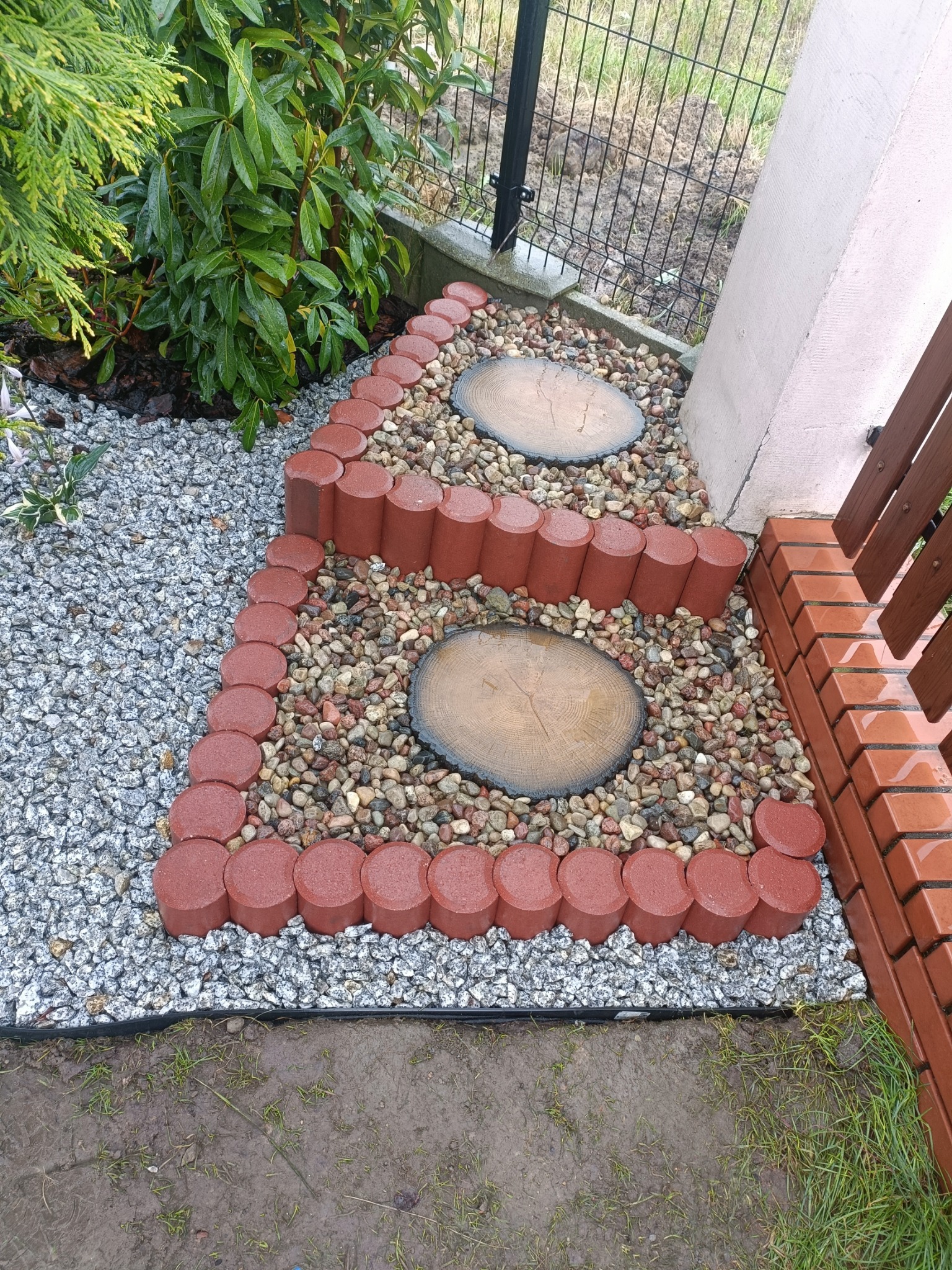 Ułożona ścieżka ogrodowa z czerwonych obrzeży, szarego żwiru i okrągłych płyt imitujących drewno, z roślinnością w tle i fragmentem ogrodzenia.