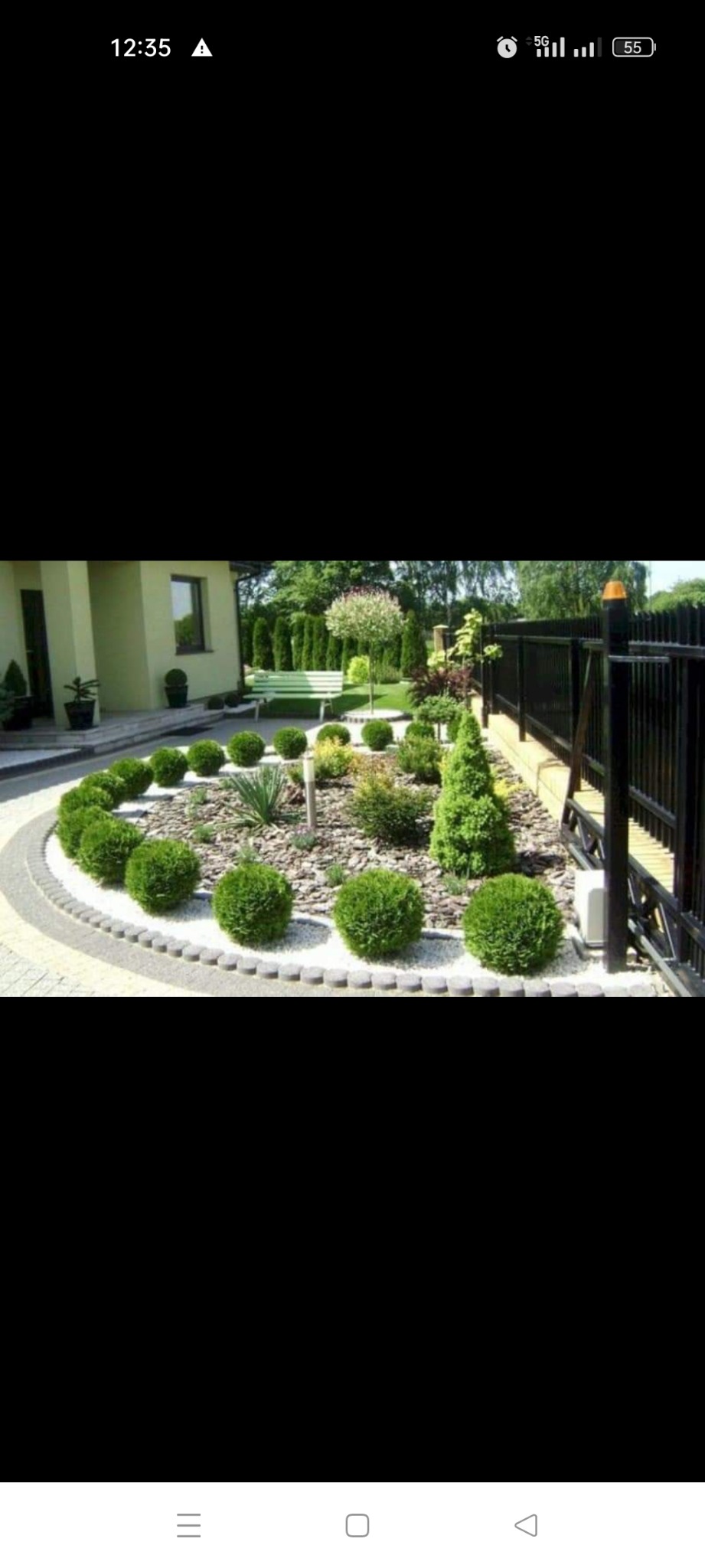 Starannie zaprojektowany i zadbany ogród przydomowy z okrągłą rabatą wysypaną białym żwirem, na której rosną kuliste krzewy, iglaki i byliny, widoczny fragment domu i czarne ogrodzenie.