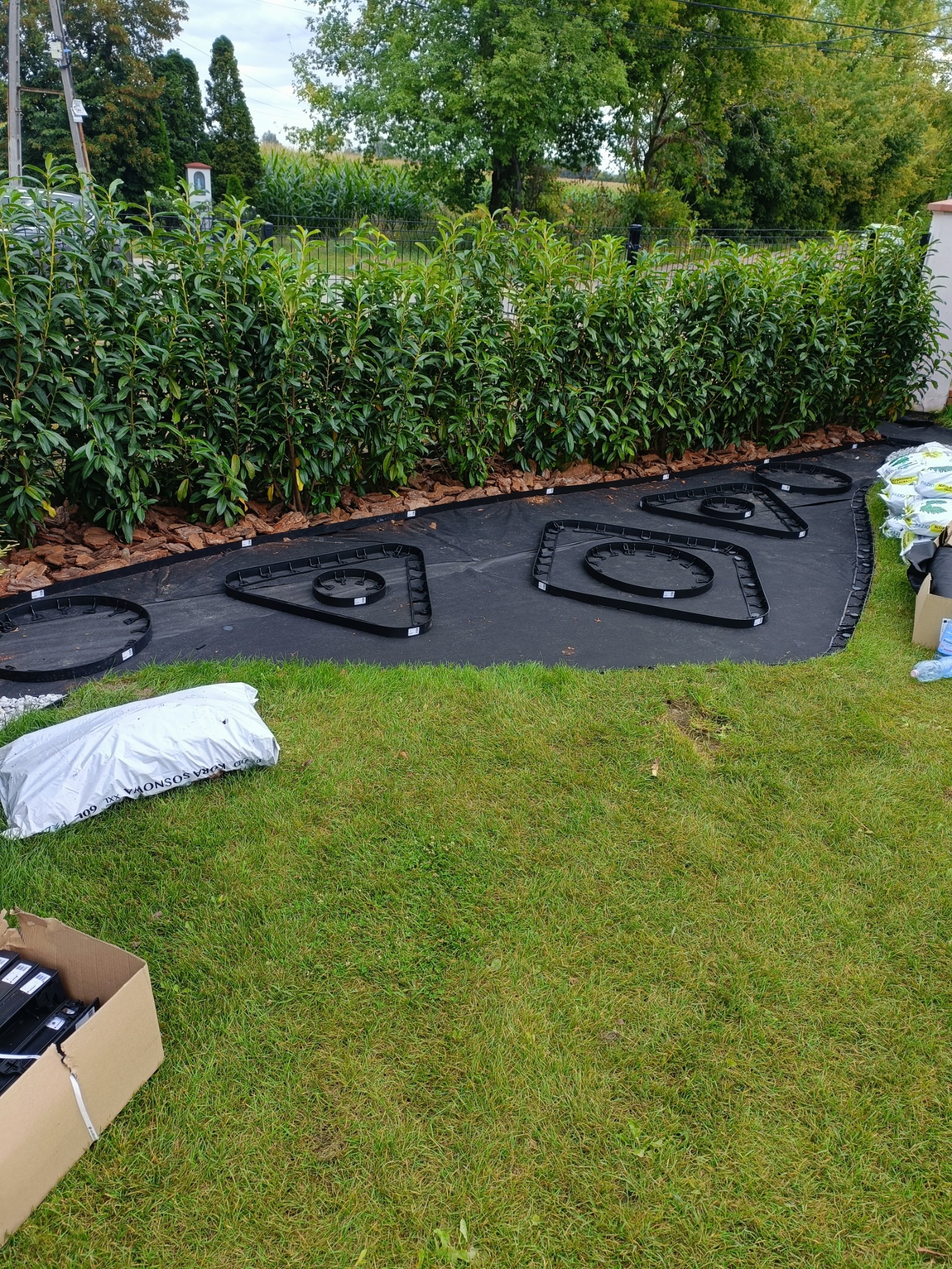 Przygotowanie podłoża pod rabaty kwiatowe: rozłożona agrowłóknina, obrzeża rabat w kształcie okręgów i trójkątów, kora sosnowa w tle, trawnik z przodu.