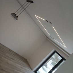 Damiam - Nowoczesne wnętrze: skośny sufit z trójkątnym oświetleniem LED, lampa wisząca i okno z widokiem na zimowe drzewa. Podłoga wyłożona panelami.