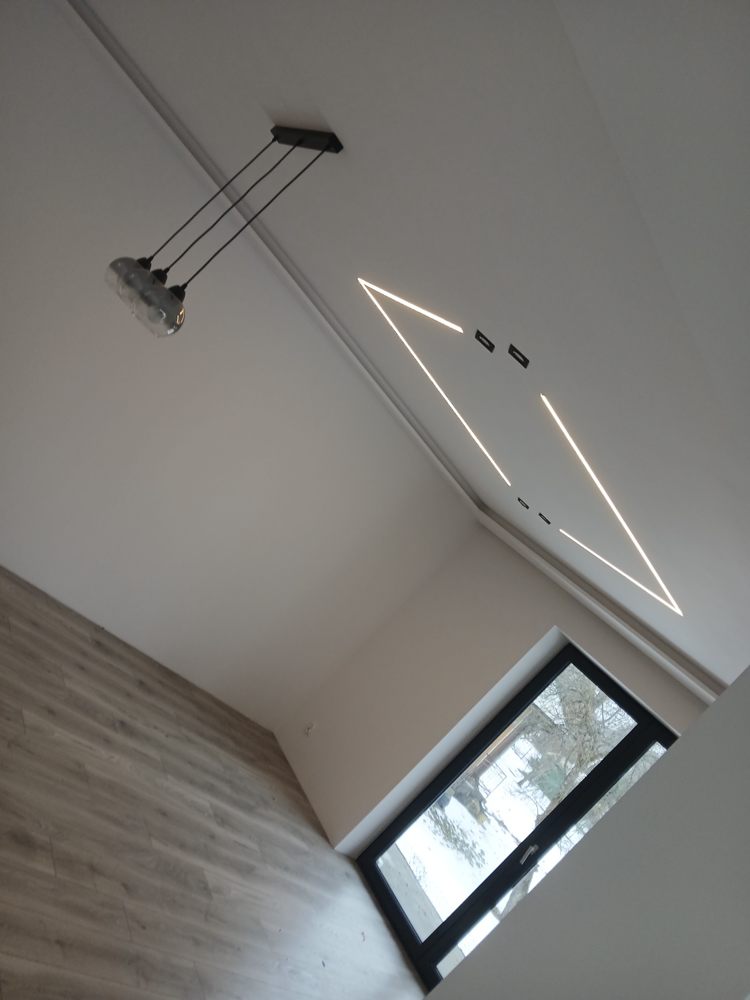 Nowoczesne wnętrze: skośny sufit z trójkątnym oświetleniem LED, lampa wisząca i okno z widokiem na zimowe drzewa. Podłoga wyłożona panelami.