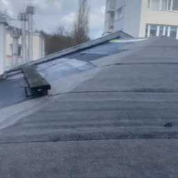 Dach pokryty papą, widoczne łatki i deska przytrzymująca fragment pokrycia, w tle budynki mieszkalne.