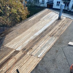 Grzegorz Zakrzewski - Widok na drewnianą konstrukcję dachu płaskiego w trakcie prac, widoczne deski, papa i narzędzie - młotek dekarski, w tle budynek mieszkalny i drzewa.