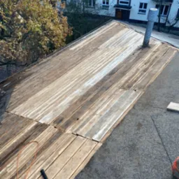 Widok na drewnianą konstrukcję dachu płaskiego w trakcie prac, widoczne deski, papa i narzędzie - młotek dekarski, w tle budynek mieszkalny i drzewa.