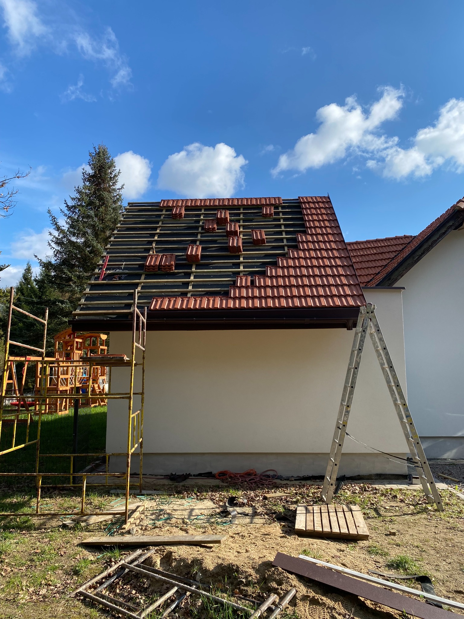 Częściowo pokryty dach z czerwonej dachówki, widoczne rusztowania i drabina, przygotowane dachówki na łatach, błękitne niebo z chmurami w tle.